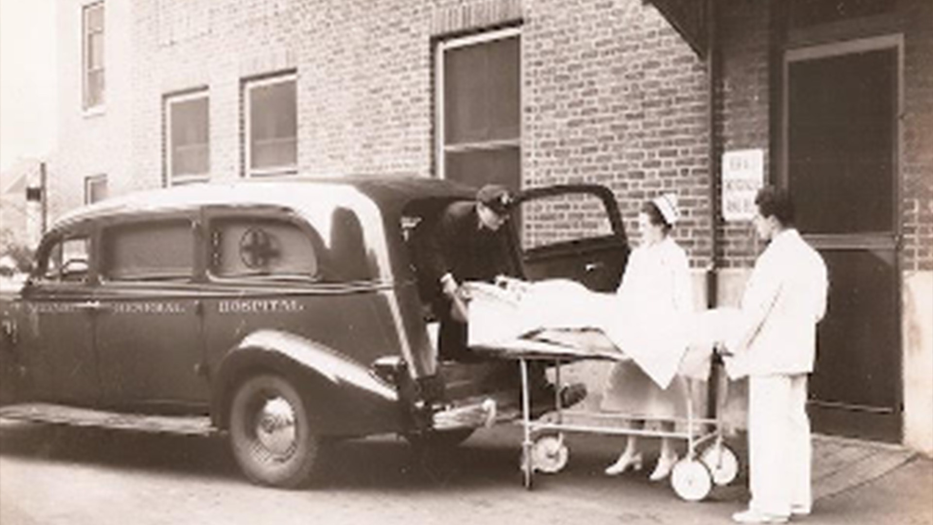 Historic black-and-white hospital scene showing staff transferring a patient from an ambulance outside an Elizabeth hospital, featured in John Mazurkiewicz’s presentation on Elizabeth’s hospital history.