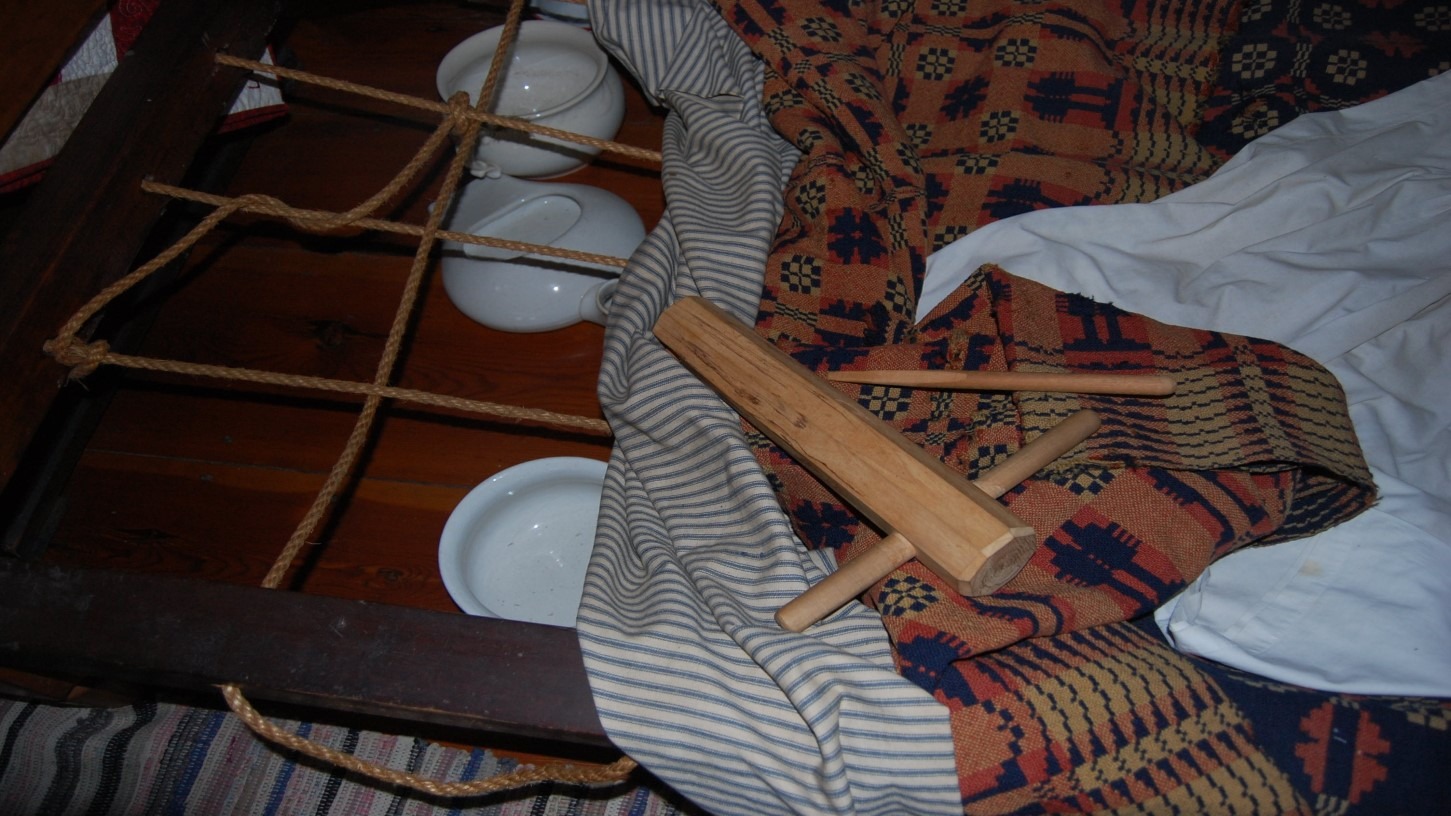 Close-up of an antique rope-strung bed with patterned coverlet, chamber pots beneath, and a wooden bed wrench.