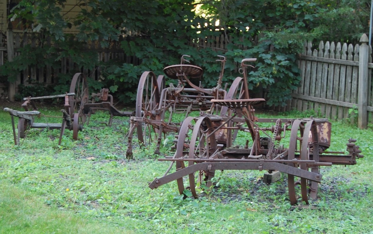 Collection of rusted antique farm machinery including plows and rakes sitting in a grassy yard