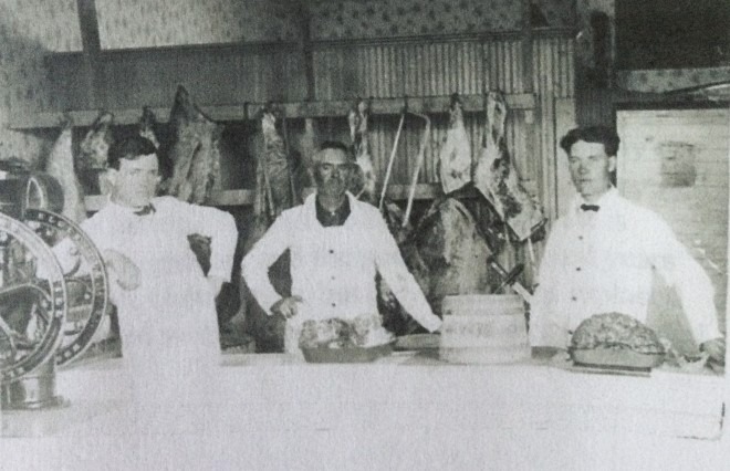 Historic photo of butchers standing behind a counter with hanging meats in a butcher shop