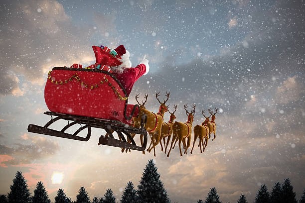 Santa Claus in a sleigh pulled by reindeer flying through a snowy Christmas Eve sky above evergreen trees